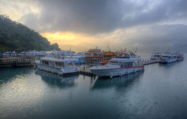 Fototapeta na wymiar Tourist boats moored to the docks during partial solar eclipse at Shuishe Pier of Sun-Moon Lake in Nantou, Taiwan, with golden sun light shining through moody cloudy sky reflected on peaceful water