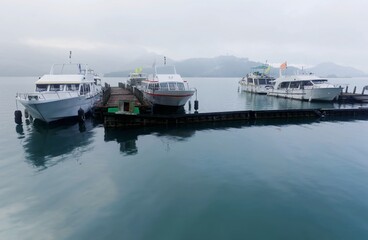 Tourist boats parking on peaceful water and moored to the floating docks of Shuishe Pier at Sun-Moon Lake on a foggy morning in Nantou, Taiwan, with mountains veiled in the fog under moody cloudy sky