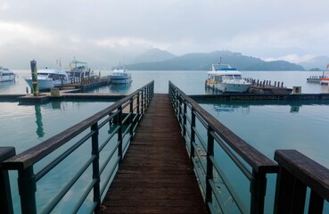 Naklejka premium Morning scenery of sightseeing boats moored to the floating docks of a wooden pier & foggy mountains by lakeside under moody cloudy sky at Sun-Moon Lake, a famous tourist destination in Nantou, Taiwan