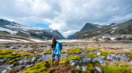 Naklejka premium Female hiker in the middle of landscape scenery overlooking Mountain peaks, icy lake and tourist cabin.
