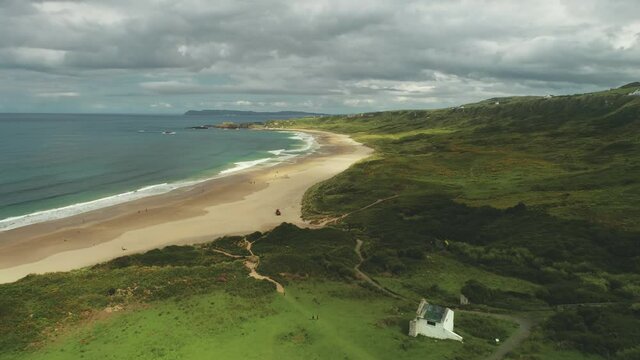 Ocean bay sandy beach aerial panoramic view under gray thick clouds. Green grass meadows valleys with white houses. People walking and looking at wonderful scenery of Irish nature. Shooting in 4K, UHD
