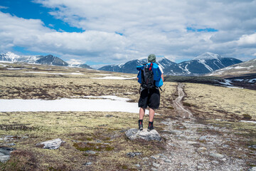 Male hiker with backpack standing on gravel path looking towards snow covered peaks during spring....