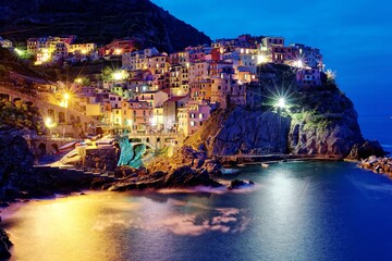 Night scenery of Manarola on vertical cliffs by the rocky coast with beautiful lights reflected on sea water, an amazing village in Cinque Terre National Park, Liguria, Italy, Europe (Long Exposure)