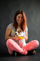 Happy smiling red-haired woman holding a small funny puppy with a yellow tulip in it mouth. The joyful owner plays with the dog Jack Russell Terrier on a black background