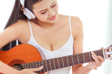 a beautiful Asian woman wake up and sitting on white bed in the bedroom, sexy girl play guitar and listen to music for relaxing in the morning