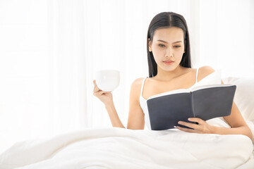 Obraz na płótnie Canvas A Asian sexy girl sitting on white bed in bedroom and drinking a cup of hot coffee with reading a book, happy vacation morning