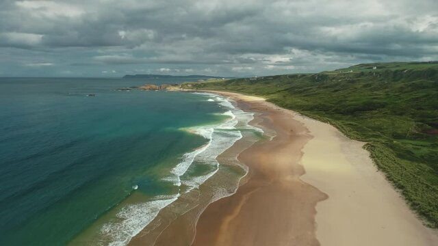 Hyperlapse aerial view: White beach gulf water crashing. People on shore walk and playing with dogs. Ocean and meadows in horizon. Picturesque beauty of Northern Ireland. Footage shooting in 4K, UHD