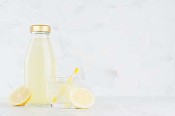 Bright yellow lemon juice in glass bottle mock up with straw, wine glass, fruit slice on white wood table in light interior, template for packaging, advertising, design, branding product.