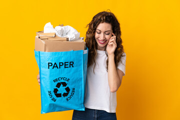 Young woman holding a recycling bag full of paper to recycle isolated on yellow background laughing