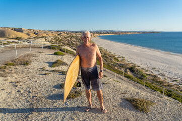 Portrait of mature senior Surfer looking at the ocean with vintage surfboard on an empty beach