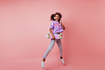 Fototapeta na wymiar Full-length shot of stunning black woman in stylish jeans jumping on pink background. Attractive curly african girl with skateboard expressing positive emotions.