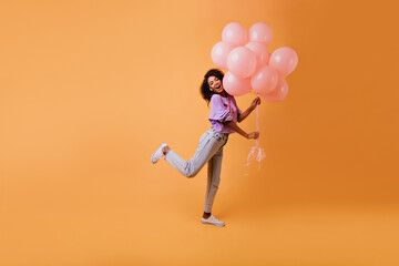 Fototapeta na wymiar Graceful african female model in casual clothes fooling around on yellow background. Indoor photo of emotional birthday girl dancing with party balloons.