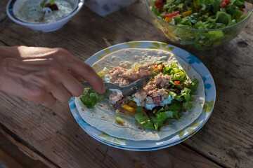 Woman's hand preparing tortilla wrap with chicken and vegetable salad, tzatziki in bowl in backround, plate on wooden table