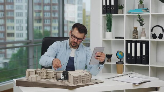 Good-looking modern serious high-skilled bearded architect in eyeglasses doing notes into i-pad during working with model of future buildings in design studio