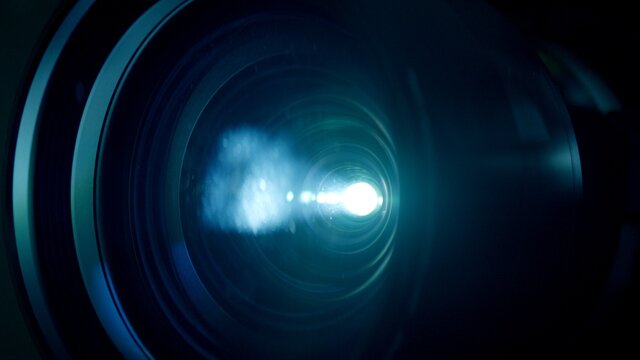 Macro closeup locked shot with selective focus of activated film projector. Illuminated lens and white light flare in dark room.