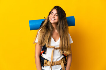 Young mountaineer woman with a big backpack isolated on yellow background looking to the side and smiling