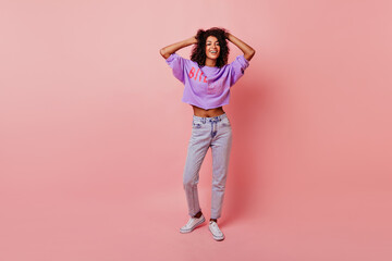 Fototapeta na wymiar Full-length portrait of lovely slim woman touching her soft black hair. Blithesome african girl in jeans dancing on rosy background.