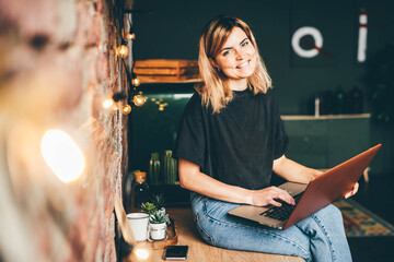 Woman in black t-shirt and denim jeans types on laptop, sitting on brown wooden kitchen table at brick wall and smiling.