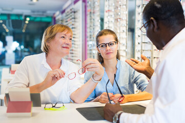 Young woman with elderly mother looking for glasses in optical shop, asking for advice from qualified African-American optometrist