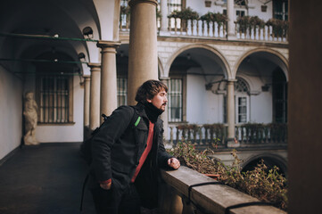 Fototapeta na wymiar A young man with long hair, a beard and mustache in a gray woolen coat, red sweater, black jeans and a backpack stands on an old balcony with baroque columns, waiting. Lviv, Italian courtyard.