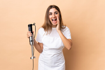 Woman using hand blender over isolated background with surprise and shocked facial expression