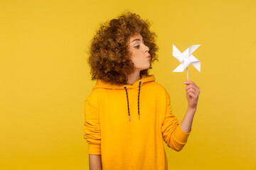 Origami hand mill. Portrait of carefree childish curly-haired woman in urban style hoodie blowing at paper windmill, playing with pinwheel toy on stick. studio shot isolated on yellow background