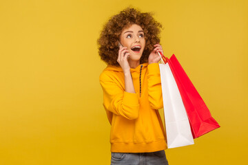 Portrait of joyful excited curly-haired hipster girl in hoodie holding shopping bags and talking on cellphone, telling friend about low prices, Black Friday. studio shot, commercial of fashion store