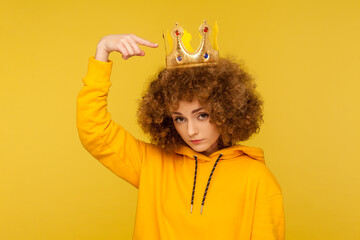 Look, I am best! Selfish haughty curly-haired woman pointing at crown on head and looking with arrogance supercilious, being egoistic with over-inflated ego. studio shot isolated on yellow background - obrazy, fototapety, plakaty