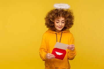 Love message. Portrait of curly-haired angelic woman with nimbus reading letter or greeting card, holding envelope, smiling and rejoicing good news. indoor studio shot isolated on yellow background