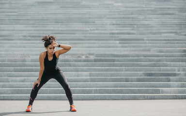 Athletic girl is tired of active training. African american young woman in sportswear with fitness tracker and wireless headphones resting on city stairs