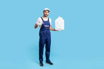 Full length professional handyman, building worker in uniform pointing at paper house, offering help in relocation, home repair services and maintenance. indoor studio shot isolated on blue background