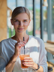 Calm beautiful happy young woman in relaxed mood looking outdoors and smiling sitting at the bar counter near big window with healthy delicious fresh lemonade glass dressed trendy white shirt