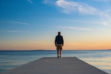 Man in hoodie and short cargo pants standing on dock and looking on sea horizon, island od Pag,...