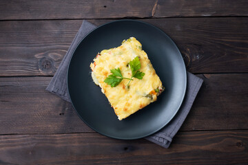 Piece casserole on a plate of potatoes and vegetables with cheese. Dark wooden background. Copy space.