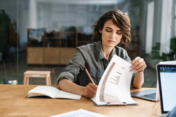 Image of young focused woman writing down notes while sitting at table - Powered by Adobe