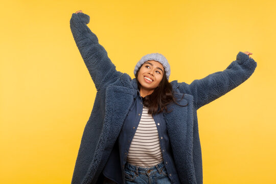 Portrait Of Carefree Cheerful Girl Wearing Warm Winter Hat And Fur Coat Raising Hands Up, Smiling Joyfully, Glamour Fashion Model In Stylish Urban Outfit Enjoying Life. Indoor Studio Shot Isolated