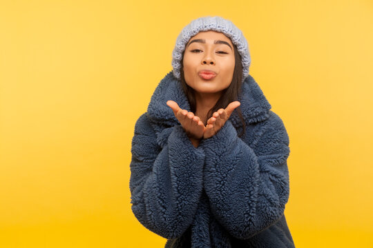 Glamour and fashion. Portrait of lovely carefree girl in warm winter hat and fur coat, stylish urban outfit, sending air kiss over palms, expressing love. studio shot isolated on yellow background