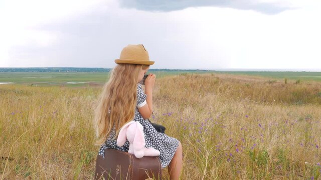 Child girl with long hair in straw hat and dress sitting on vintage suitcase and taking a picture. Cute kid with soft toy looking at nature lanscape background. Adventure travel concept in retro style