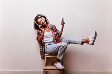 Graceful young woman in blue jeans listening music on white background. Indoor shot of carefree african girl in headphones smiling with eyes closed.