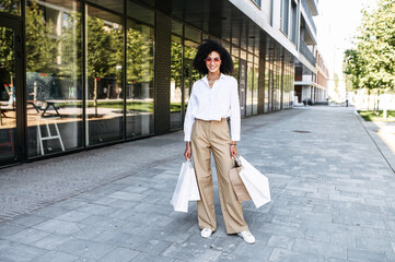 A biracial girl satisfied with shopping walks around town. A young African woman in a stylish wear holds many paper shopping bags in hands and looks at the camera