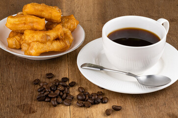 Normal breakfast with deep-fried dough stick in a white ceramic plate, pile of coffee beans hot black coffee in a white ceramic cup and metal spoon on wooden table.