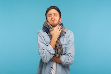 Portrait of unhealthy flu-sick man standing wrapped in warm scarf shivering from cold, feeling unwell suffering fever, seasonal influenza symptoms. indoor studio shot isolated on blue background