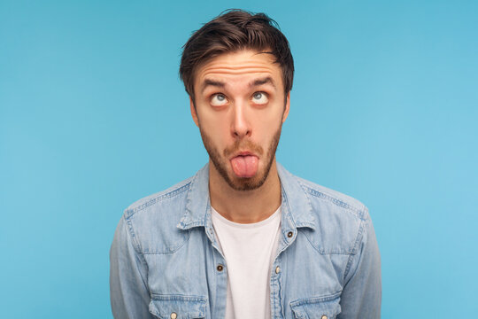Portrait of idiot man in denim shirt making silly humorous face with eyes crossed and tongue out, showing comical silly brainless facial expression. indoor studio shot isolated on blue background