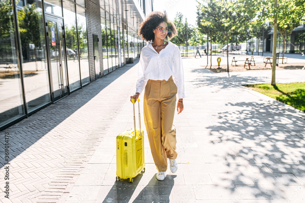 Wall mural attractive trendy young woman going to travel. a girl with an afro hairstyle with a yellow suitcase 