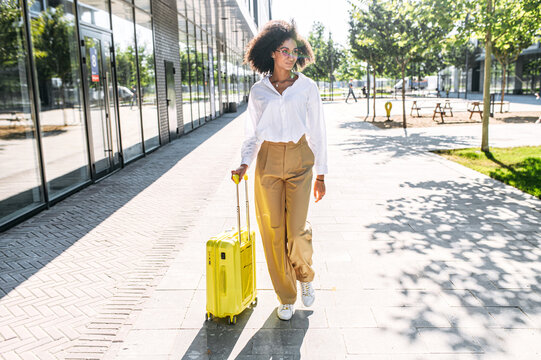 Attractive trendy young woman going to travel. A girl with an Afro hairstyle with a yellow suitcase walks on the street to station, airport. Full length