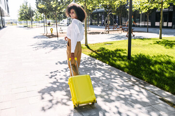 A trendy multi-ethnic woman walks with a trolley bag outdoors. A flirty girl with a suitcase looks...