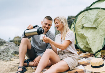 Young couple travelers in the casual outfits with tent. Man pours tea from thermos. Local tourism, weekend trip