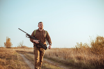 Strong young hunter with red beard holding his gun and walking along the dirt road under blue sky