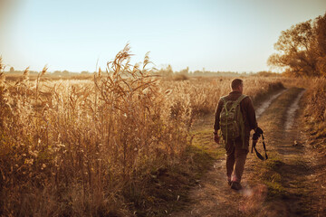 Strong young hunter with red beard holding his gun and walking along the dirt road under blue sky