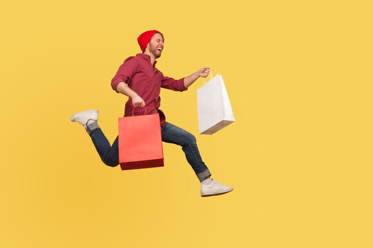Enthusiastic joyful hipster trendy guy running in air with shopping bags in hands, hurrying to catch sale, flying and rushing for thrift discount. full length studio shot isolated on yellow background